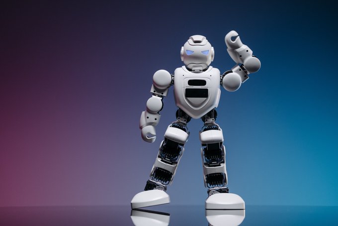 The image features a white robot with blue eyes, standing upright against a purple background. The robot appears to be interacting with its surroundings or posing for a photo. The caption above the image provides information about the robot's creator and their approach to AI development.
