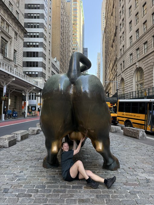 A photograph of a person sitting on cobblestone pavement beneath the rear view of the 'Charging Bull' sculpture in New York's Financial District, with a yellow school bus and tall buildings in the background.