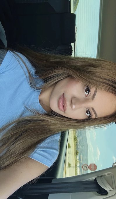 A young woman with long brown hair wearing a light blue t-shirt takes a close-up selfie inside a car, with a scenic view visible through the window. The image pairs with the cheerful "gm cuties !" greeting, presenting a fresh morning look.