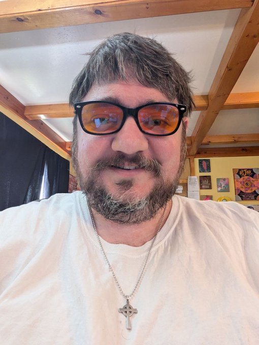 A man with a beard and reflective sunglasses smiles warmly in a selfie, wearing a white t-shirt and a silver cross necklace, with framed photos and posters visible in the background. The image conveys authenticity and approachability, aligning with the post's message about the importance of being genuine as a Bitcoiner.