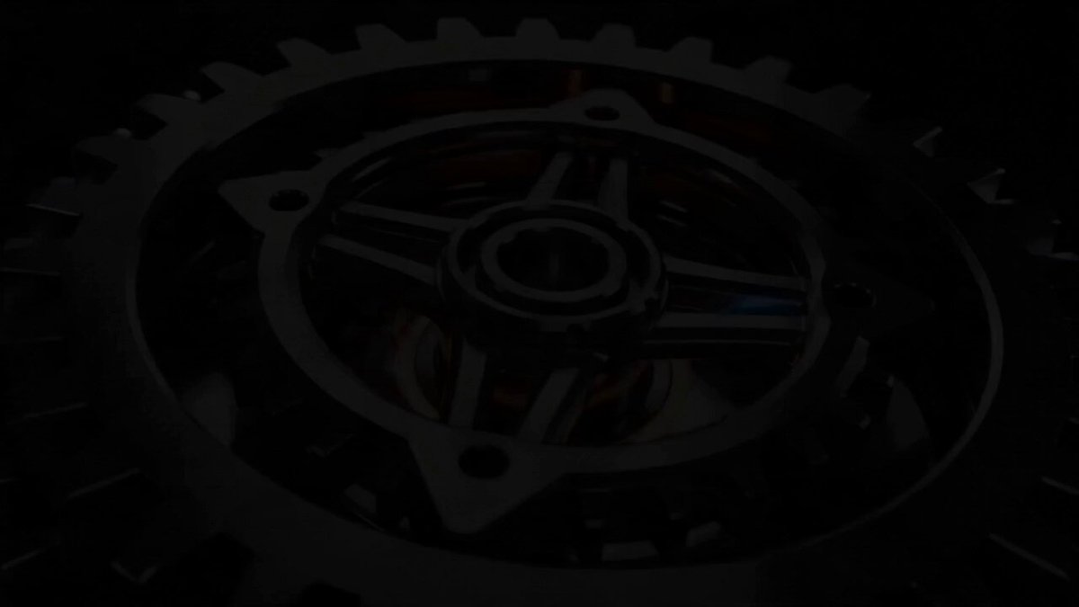 The image shows a close-up view of a gear wheel, which appears to be black in color. The gear wheel has a unique design with multiple teeth, giving it an industrial appearance. The background of the image is dark, making the gear wheel stand out even more. There are no texts or other objects present in the image, and the focus is solely on the gear wheel.