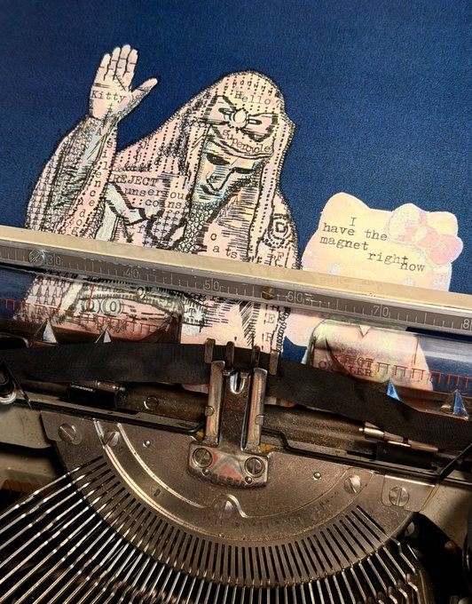 A vintage typewriter sits in front of a blue background featuring artwork of a hooded figure composed of text and collage elements, with a small label reading 'I have the magnet right now'.