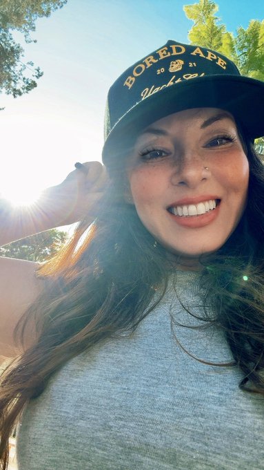 The image features a woman wearing a black baseball cap with "Bored Ape" written on it, smiling at the camera while posing for a photo. The caption below the photo reads, "I am a Bored Ape. I am part of the best Club on the internet. I celebrate the community we've built. I am proud to be a member of the Club."