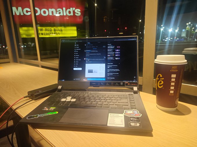 A laptop sits open on a McDonald's table next to a coffee cup, with the illuminated McDonald's sign visible through the window at night.