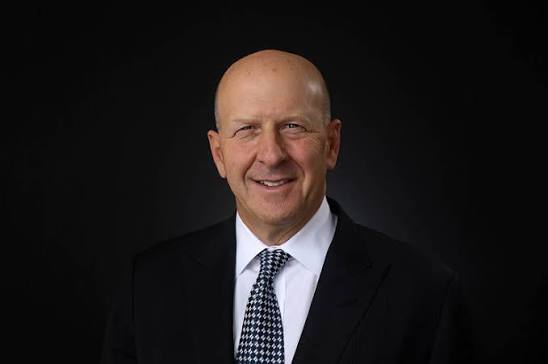 A professional headshot of a smiling, bald man wearing a dark suit, white shirt, and a patterned tie, photographed against a black background.