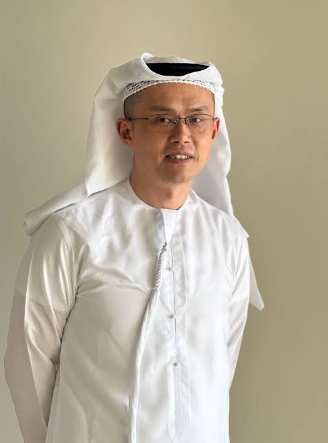 A portrait of a man wearing glasses and traditional Middle Eastern attire, including a white kandura and a white ghutra headdress with a black agal, smiling slightly against a plain beige background.