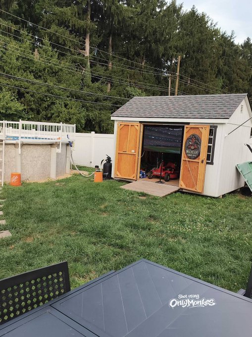 A backyard scene showing a wooden shed with open doors revealing a red riding lawnmower inside, set on a green lawn with a white fence, above-ground pool, and tall trees in the background.