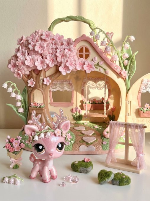 A pink Littlest Pet Shop-style deer figurine wearing a floral crown stands in front of an elaborate blossom-themed dollhouse featuring a cherry blossom tree, flower-covered details, and a swing set, capturing a whimsical, fairytale-like scene.