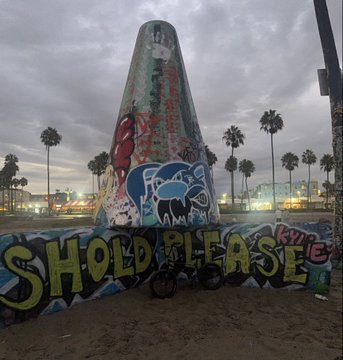 A graffiti-covered concrete cone monument with 'SHOULD PLEASE' painted at the base, set against a cloudy sky with palm trees.