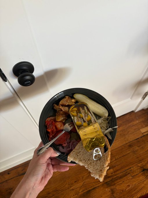 A first-person view of someone holding a bowl of food including a sandwich, pickles, onions, and a pull-tab can, standing near a white door with a barbell on the floor.