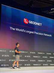 A man presenting on stage at a GEODNET event, with a large screen displaying 'GEODNET - The World's Largest Precision Network' and logos for partners including Bitcoin and Decentral.
