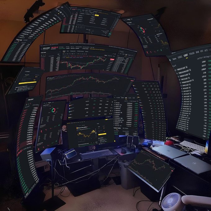An elaborate multi-monitor trading setup displaying various Bitcoin and cryptocurrency charts, candlestick graphs, and ticker listings on numerous screens arranged in an arc.