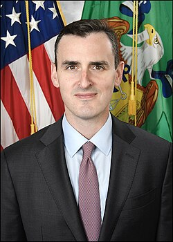 The image shows a man in a dark suit and pink tie posing in an official portrait with the American flag and a green government seal in the background; the accompanying post discusses IMF official Dan Katz's warnings about local-currency stablecoins accelerating dollarization, suggesting the man pictured may be the official referenced in the article.