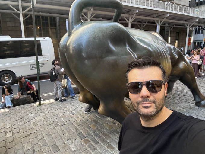 A selfie of a man in sunglasses in front of the Charging Bull statue in New York City's Financial District, with a white tour bus and pedestrians in the background.