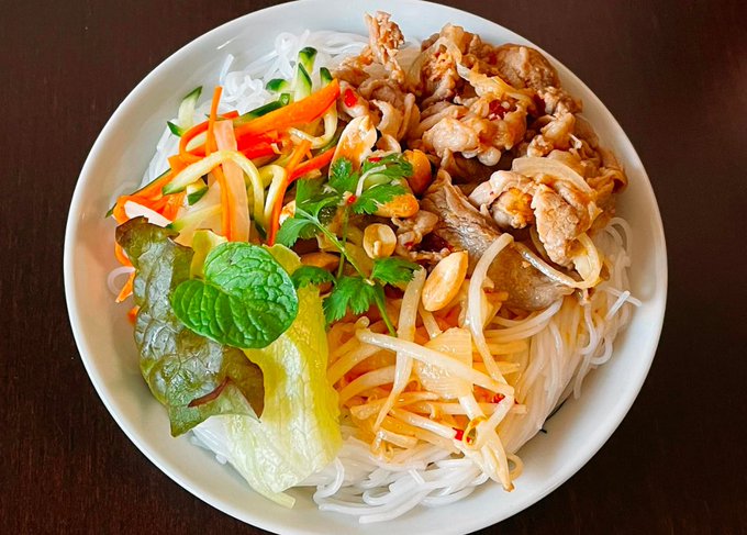 A white bowl filled with rice noodles topped with grilled meat, sliced cucumbers, shredded carrots, fresh herbs, peanuts, and lettuce.