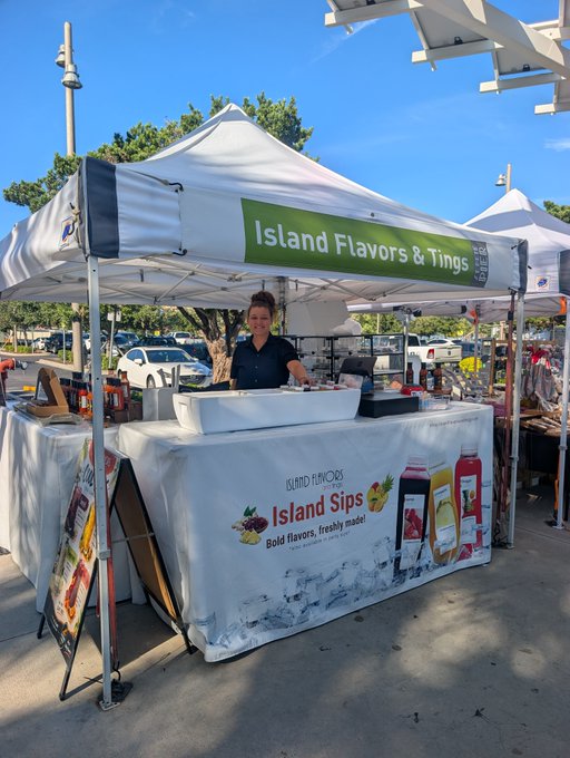 A vendor stands under a white tent with a green banner reading 'Island Flavors & Tings', with a table banner advertising 'Island Sips' and 'Bold flavors, freshly made'.