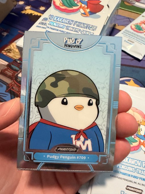 A hand holding a holographic 'Pudgy Penguins' trading card featuring a cartoon penguin character wearing a camouflage helmet, labeled 'Pudgy Penguin #709 Phantom'.