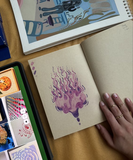 A hand with gold rings rests on an open kraft paper sketchbook featuring a vibrant purple and pink flame-like illustration, with additional artwork visible in surrounding notebooks and a tablet. The "Should I mint it?" caption suggests the artist is considering turning their striking purple flame design into an NFT.