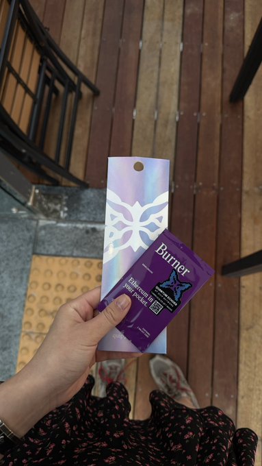 A hand holds a purple 'Burrier' pain relief patch packet and its patterned cardboard packaging while standing on wooden decking.