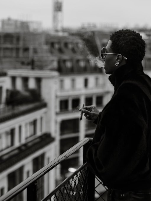 A black-and-white photograph of a woman in sunglasses and a dark coat smoking a cigarette on a balcony overlooking a European city skyline.