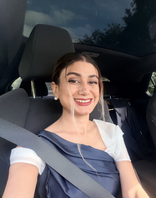 A smiling woman with brown hair wearing a navy blue and white top sits in the driver's seat of a car, secured with a seatbelt.