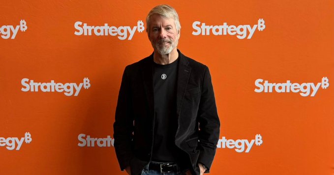 A man stands confidently in front of an orange backdrop repeatedly displaying the "Strategy" logo, representing the company that just resumed its $BTC buying spree with a $369.7M acquisition, bringing its total holdings to 845,050 BTC.