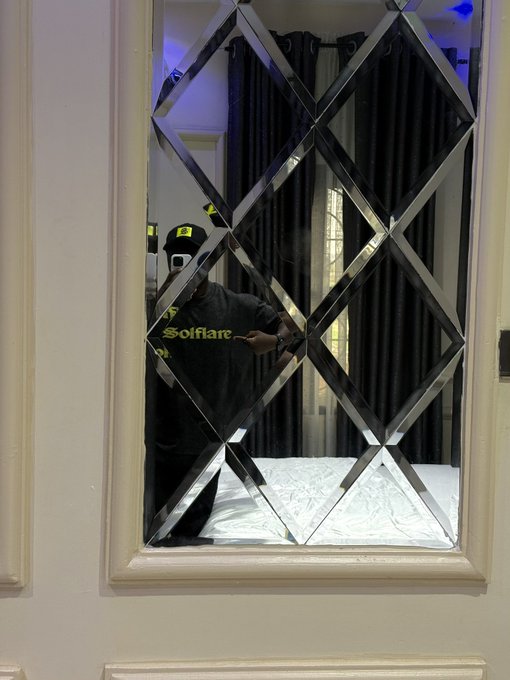 A person wearing a dark shirt with 'Solfare' in yellow lettering taking a mirror selfie in a decorative diamond-patterned mirror, with a bed visible in the reflection.
