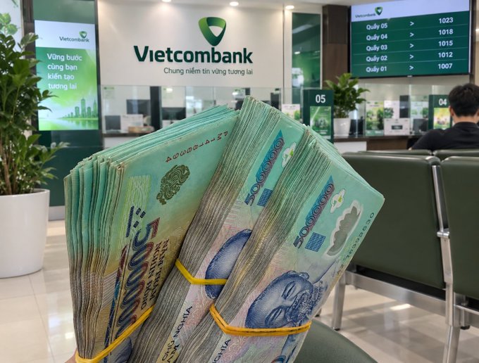 A hand displays thick stacks of 50,000 Vietnamese Dong banknotes secured with yellow bands inside a Vietcombank branch, with the bank's logo and the slogan 'Vững bước cùng bạn kiến tạo tương lai' visible in the background.