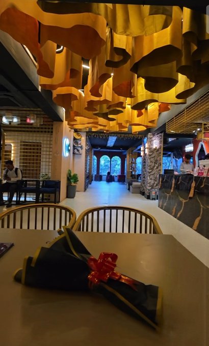The interior of a restaurant or cafe featuring distinctive golden, wavy hanging ceiling decorations, with a wrapped gift with a red bow sitting on a table in the foreground.