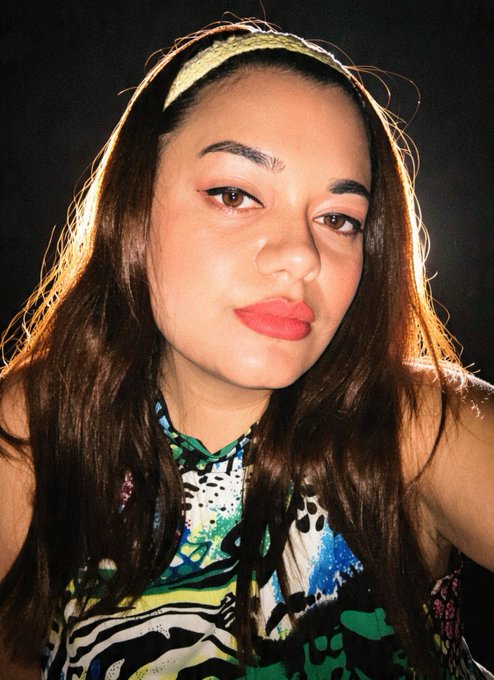 A close-up portrait of a young woman with long dark hair wearing a patterned sleeveless top and a headband, with red lipstick and a neutral expression.