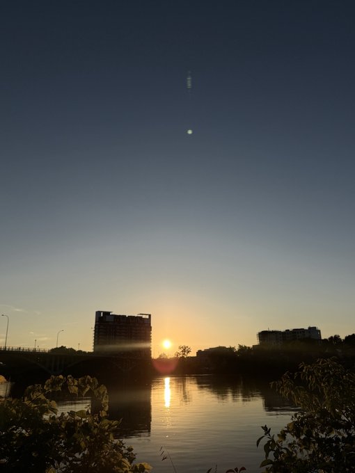A serene sunset photo over a river with city buildings silhouetted on the horizon, featuring a bright sun reflection on the water and an unidentified bright object high in the evening sky.