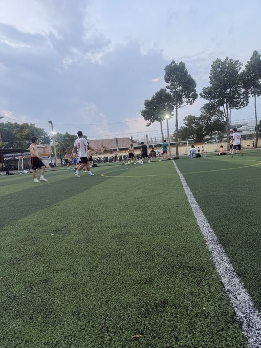 People play soccer on a green artificial turf field in the late afternoon, with trees, stadium seating, and a cloudy sky visible in the background.