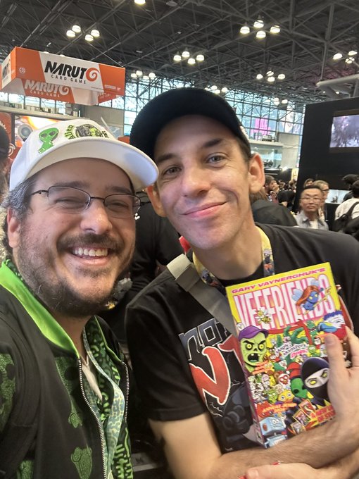 Two men posing together at what appears to be a gaming convention (Naruto booth visible), one wearing a cap and glasses with a beard, the other wearing a black cap and shirt, holding a Gary Vay-Neerchuk's 'VFRIENDS' book/comic.