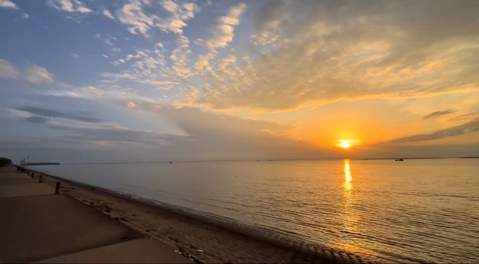 A scenic sunset over a calm body of water with the sun reflecting on the surface, dramatic clouds overhead, and a stone seawall along a shoreline.