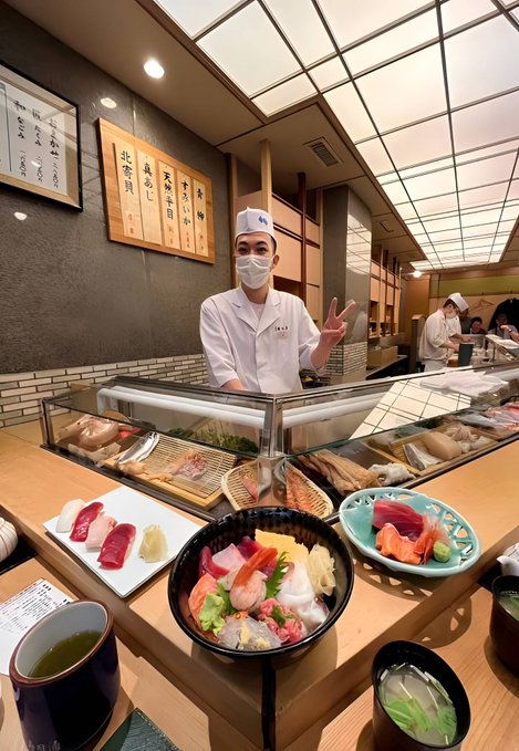 A smiling sushi chef wearing a white uniform, hat, and mask gives a peace sign behind a counter displaying an assortment of fresh sashimi, nigiri, and chirashi bowls, with other kitchen staff visible in the background. The playful caption "GM - he says hi too" humorously matches the chef's friendly peace sign greeting alongside the morning scene.