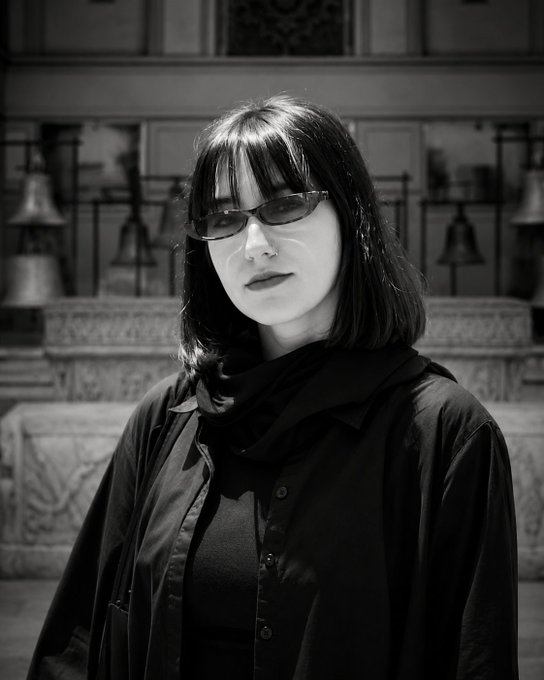 A black and white portrait of a woman with dark hair, bangs, sunglasses, and a nose ring wearing a dark shirt and scarf.