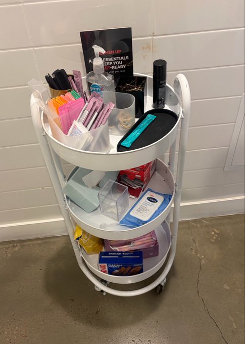 A three-tiered white rolling cart filled with various toiletries, stationery, cleaning supplies, and personal care items against a white tiled wall.
