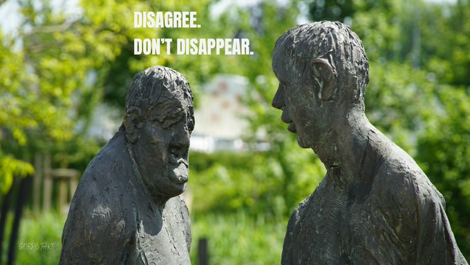 Two dark sculptures of figures face each other in front of greenery, with text reading 'DISAGREE. DON'T DISAPPEAR.'