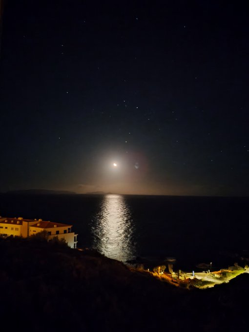 a nighttime view of the ocean with the moon reflecting on the water, a few stars visible in the sky, and buildings lit up on the shore.