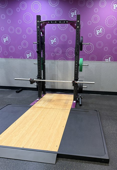 A gym setup featuring a power rack with a barbell and green weight plates on a wooden lifting platform, with a purple wall branded with 'pf' (Planet Fitness) logos in the background.