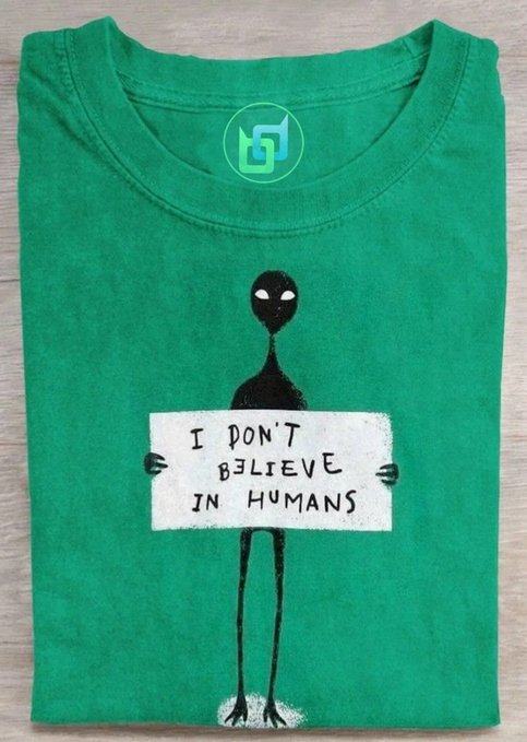 A folded green t-shirt features a black alien graphic holding a sign reading "I DON'T BELIEVE IN HUMANS." The image ties into the post's theme of feeling misunderstood, using the alien's distrustful message to humorously represent the Web3 perspective of $BDX (Beldex Coin) holders who see crypto's potential while others remain skeptical.