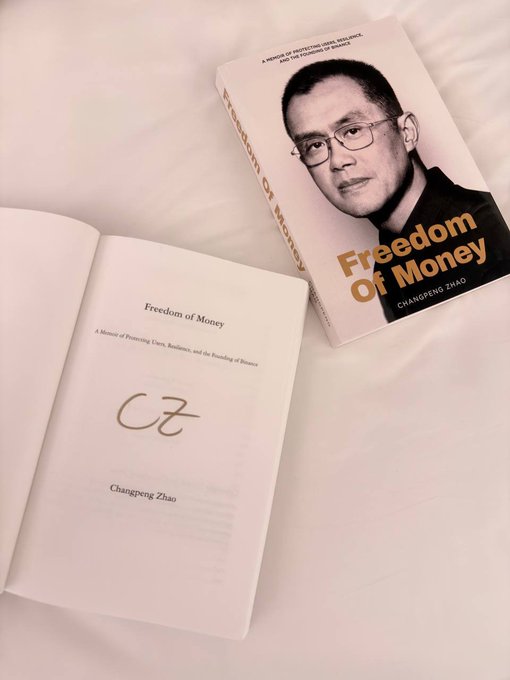 A signed copy of "Freedom of Money" by Changpeng Zhao (CZ) rests on a light surface, with its dust jacket displaying a portrait of the author pulled aside to reveal the title page inside. The book personally signed by CZ serves as a meaningful memento from the Binance Campus Asia 2026 event, making it a treasured keepsake for the recipient.