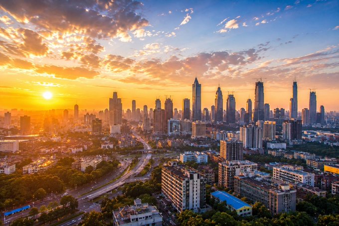 A sprawling city skyline glows beneath a vibrant sunrise, with skyscrapers silhouetted against golden clouds as morning traffic flows through the urban landscape below. The sunrise imagery reinforces the "Good Morning, CT" greeting and the optimistic TGIF message, symbolizing a fresh start and growth—mirroring the post's theme of building wealth through XORA's AI-driven yield deposits.
