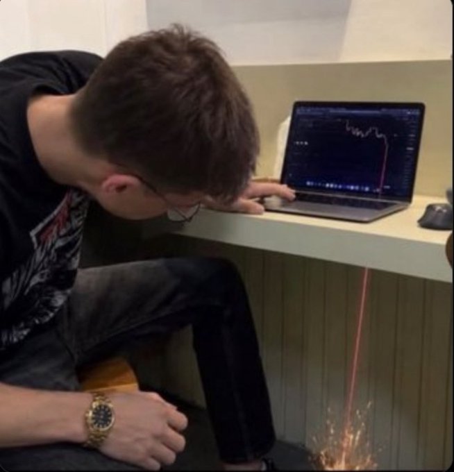 A young man in a black t-shirt leans his head against a desk in apparent defeat as a stock chart on his laptop shows a dramatic downward crash with a red line plummeting.