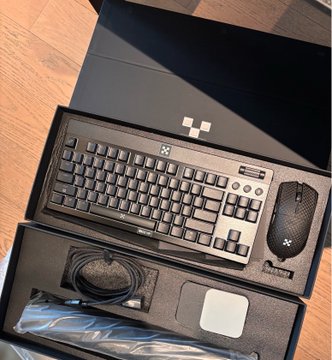 An open black box on a wooden floor containing a Razer keyboard, mouse, and a white square item, with cables in packaging.
