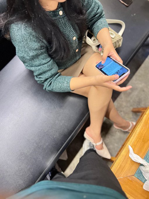 A person sits on a gray bench wearing a green knit top and beige skirt, holding a smartphone with both hands.