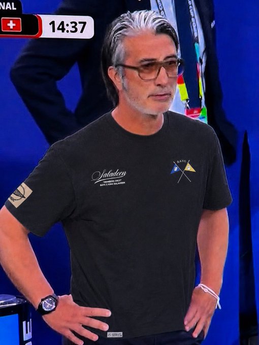 The image shows a man with grey hair and glasses wearing a black "Salaten" t-shirt with crossed flags emblem, standing at what appears to be a sporting event with a Swiss flag and timer showing 14:37 in the background. The "bored ape yacht club" reference appears to relate to BAYC branding visible on the lanyard behind him, suggesting a connection to the NFT community at the event.