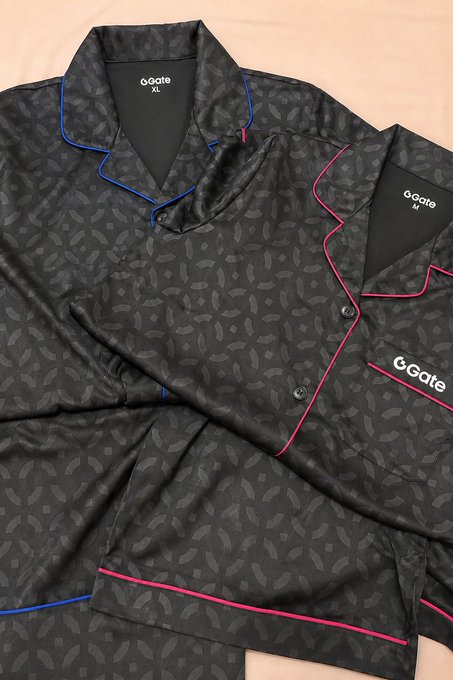 Two black patterned pajama sets laid out on a pink surface, branded 'Goate' with one labeled size XL (blue piping) and the other size M (pink piping).