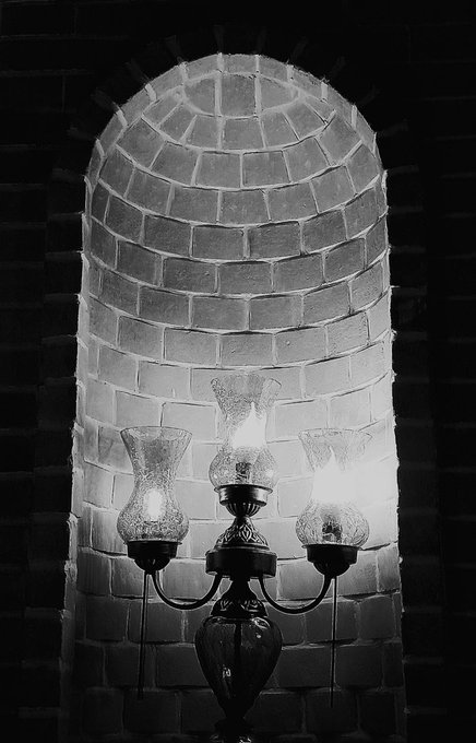 The image features a four-sided lantern sitting inside an arched doorway, with light shining through it onto the brick wall behind it. The interior of the room looks dark and shadowy. Above the doorway, there is some text that reads "Happy #TEZOSTUSDAY Light in the Darkness". The overall atmosphere appears to be dimly lit or darkened, contrasting with the warm glow coming from the lantern's light source.