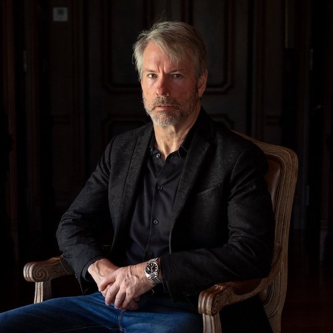 A silver-haired man in a dark blazer and black shirt sits in an ornate wooden armchair, gazing directly at the camera with a composed expression. The portrait accompanies news of Strategy's (formerly MicroStrategy) $369 million Bitcoin purchase of 4,603 BTC, reportedly acquired after selling at a cycle low—a move tied to founder Michael Saylor's well-known advocacy for Bitcoin as a corporate treasury asset.
